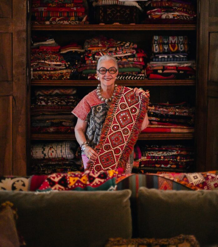 Textile collector Suzanne Sugg displaying a vibrant hand-embroidered heritage textile in front of her extensive global fabric collection.