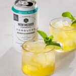 Two glasses of a yellow cocktail with a sprig of mint sit in front of a can of New Mexico kombucha.