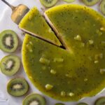 A black sesame kiwi cheesecake with a gelée layer in green sits witha. slice being taken out and kiwi slices all around the outside.