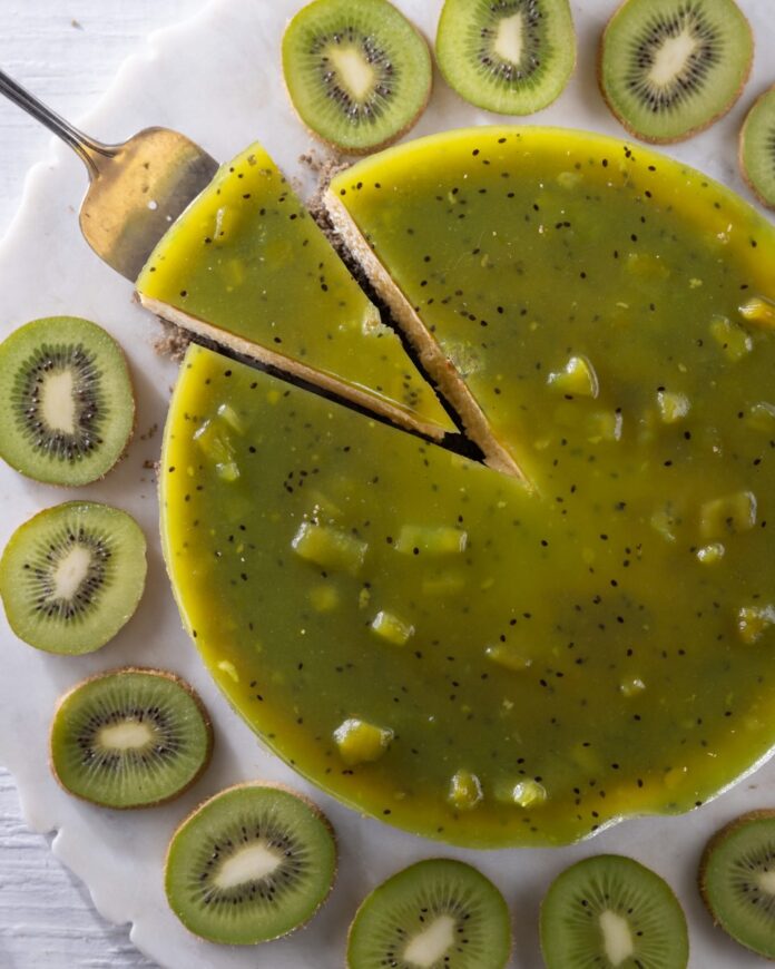 A black sesame kiwi cheesecake with a gelée layer in green sits witha. slice being taken out and kiwi slices all around the outside.
