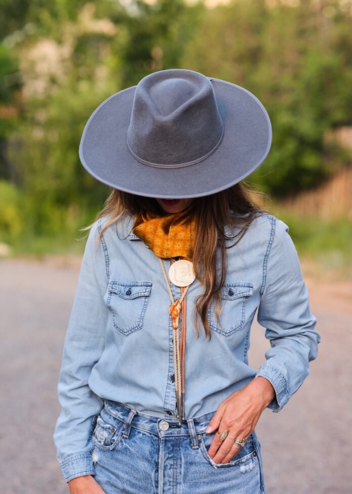 A woman in a blue hat looks down at her blue jean shirt and pants with an orange bolo tie.