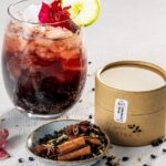 An elderberry spritzer sits on a white table with a lime wheel and flower in the glass, elderberry syrup kit beside it, and cinnamon sticks in a small bowl in front of it.