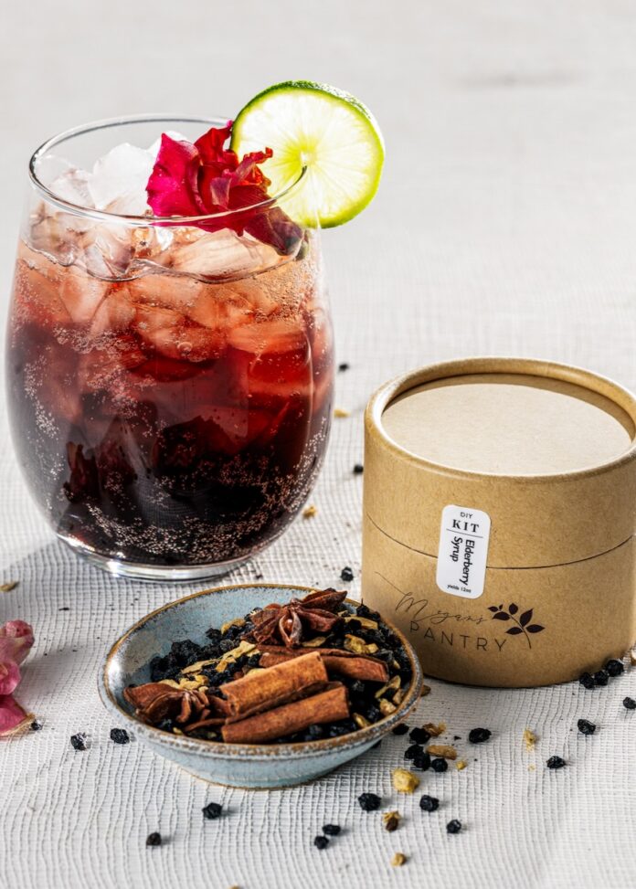 An elderberry spritzer sits on a white table with a lime wheel and flower in the glass, elderberry syrup kit beside it, and cinnamon sticks in a small bowl in front of it.