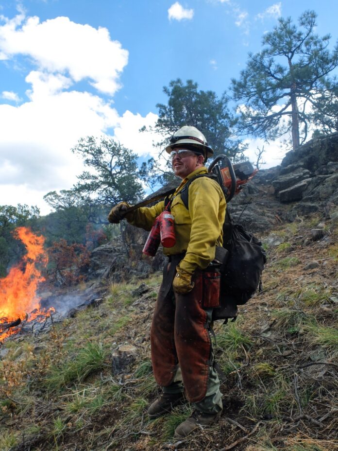 A man stands in fire gear with a forest wildfire burning in the background.