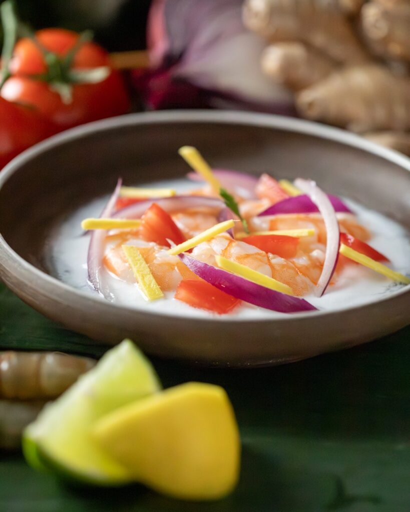 A small bowl of shrimp ceviche in a white coconut milk next to two slices of citrus and with slices veggies as a garnish.