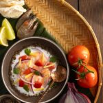 A small bowl of Kinilaw na Hipon (Filipino Ceviche) sits with a spoon in it on a woven tray next to tomatoes and citrus.