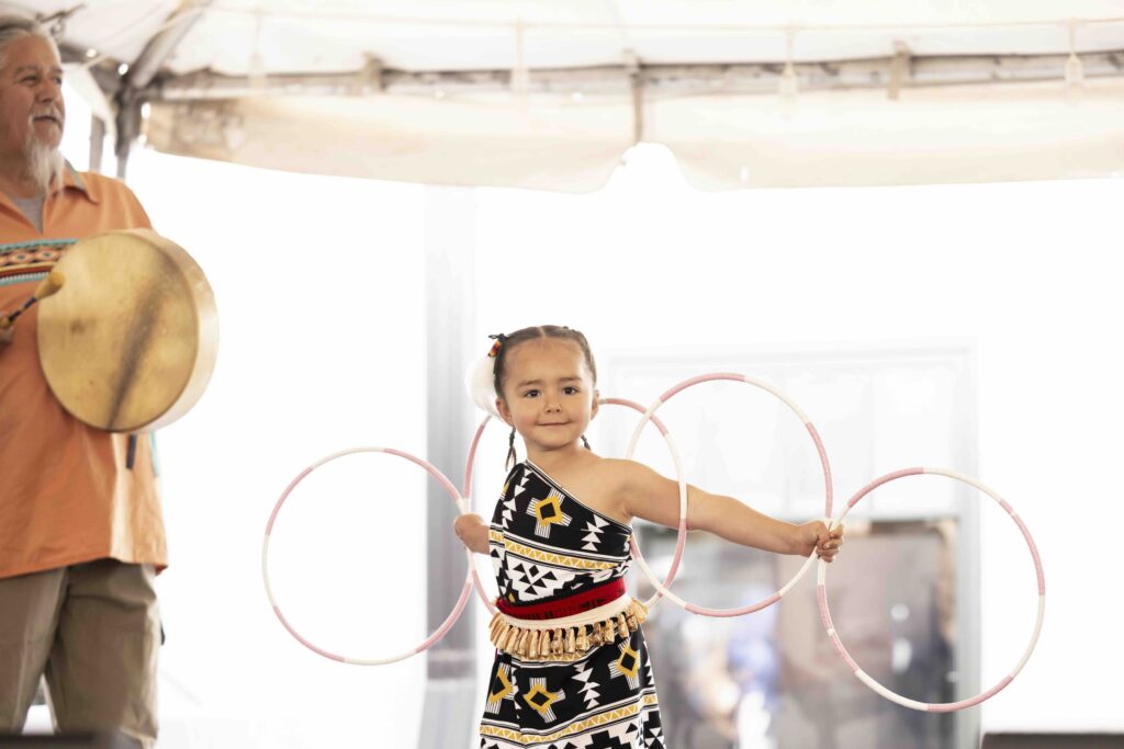 A young Native American girl with mini rings used for dancing and an older gentleman with a hand drum.