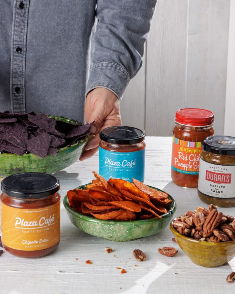 A man moves a bowl of chips on the table that sit around various jars of salsas.