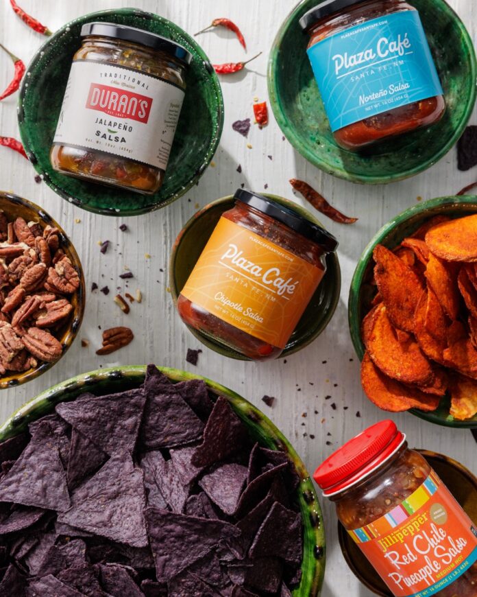 A variety of New Mexico made salsas in jars that sit in small green bowls around bowls of chips.