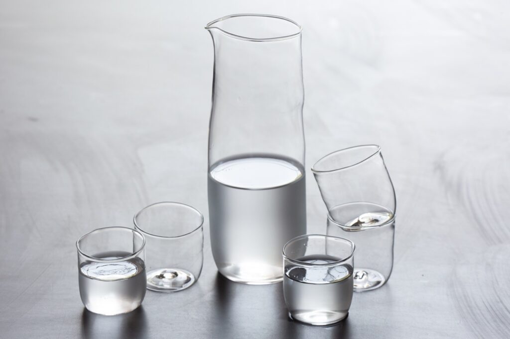 Five small drinking glasses beside a larger glass pitcher.