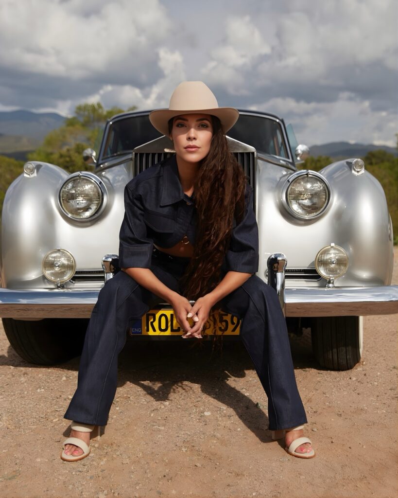 A woman in a brimmed hat sits on the front bumper of a silver antique car. 