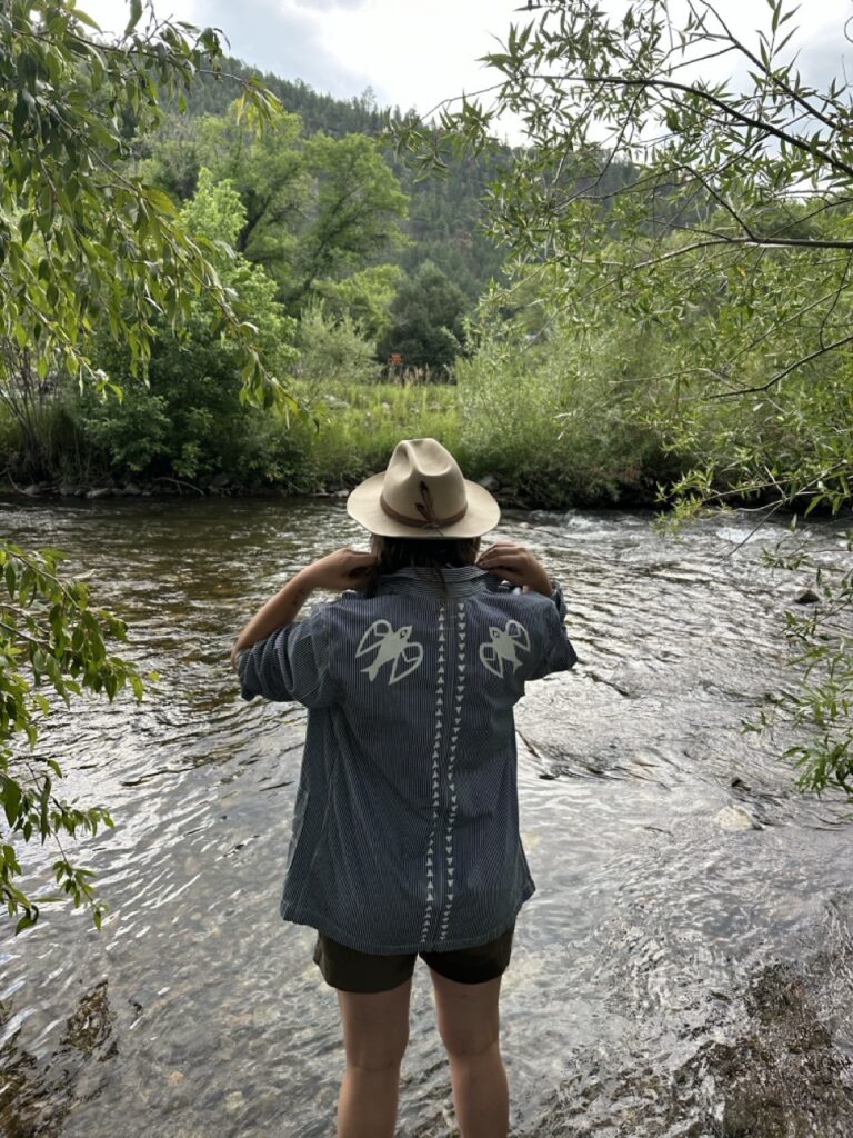 A woman in a collar shirt and brown hat stands in a creek.