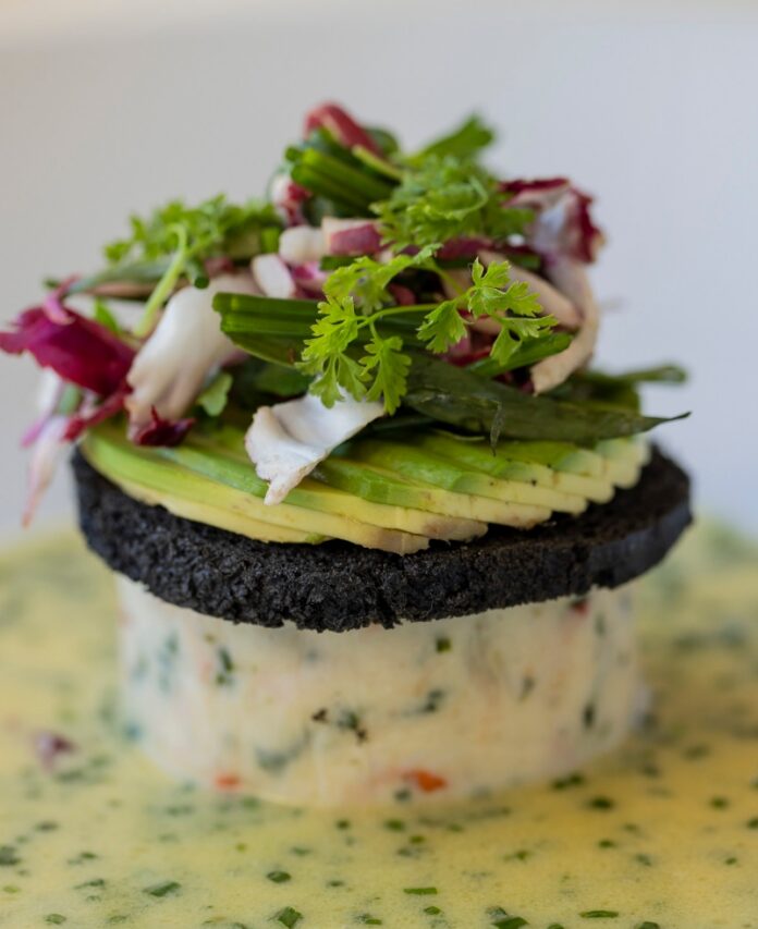 A gourmet tower of Maine lobster and bay scallop mousse topped with black squid ink brioche, sliced avocado, and fresh greens in a chive beurre blanc sauce at The Compound restaurant in Santa Fe.