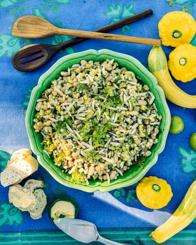 A vibrant summer grain salad with corn, beans, and herbs served in a green bowl alongside fresh yellow squash.