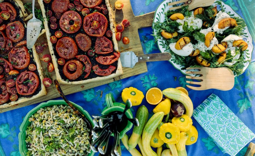 A tailgate feast featuring tomato and tapenade tarts, caprese salad with peaches, and fresh summer vegetables.