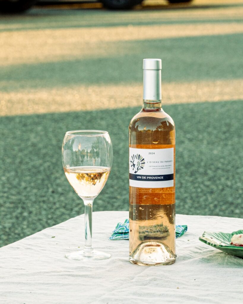 A bottle of L’Oiseau du Paradis Rosé 2024 and a filled wine glass on a white tablecloth during a sunset tailgate.