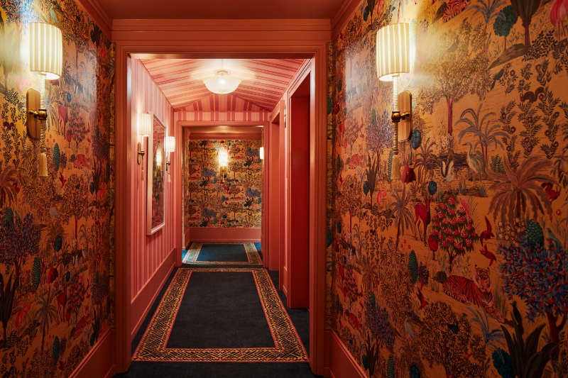 A vibrant hotel corridor at The Fifth Avenue Hotel with pink walls, tropical patterned wallpaper, and a blue bordered carpet leading to guest rooms.