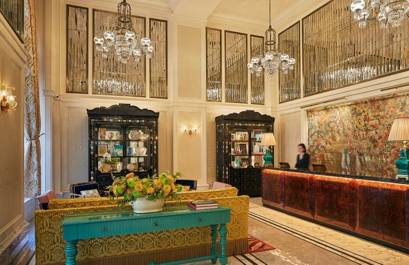 The dazzling lobby of The Fifth Avenue Hotel featuring burled wood panels, faceted mirrors, and an artistic tapestry by Pae White.