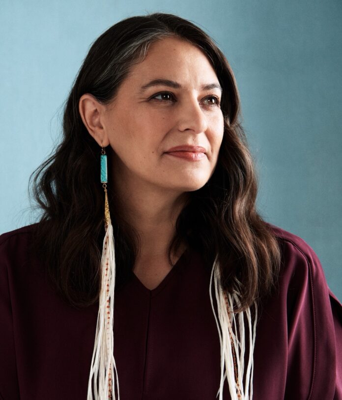 Candice Hopkins stands against a blue background with Indigenous style earrings hanging down with feathers and turquoise.