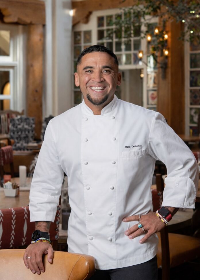 Chef Marc Quinones in a white chefs jacket with his hand on the back of a wooden chair.