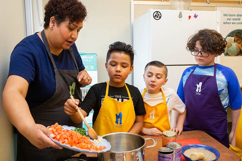Three kids and a teacher cooking by putting ingredients into a pot.