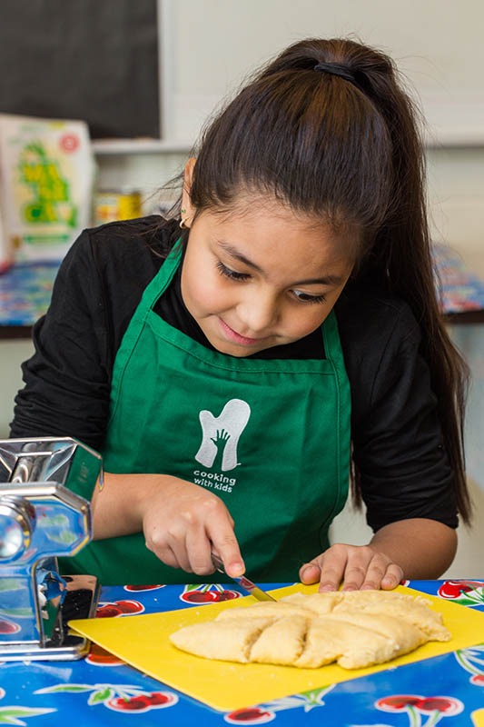 Meet Carmen Benson, Executive Director of Santa Fe's Cooking with Kids A little girl chopping up dough on a cutting board with a green apron on.