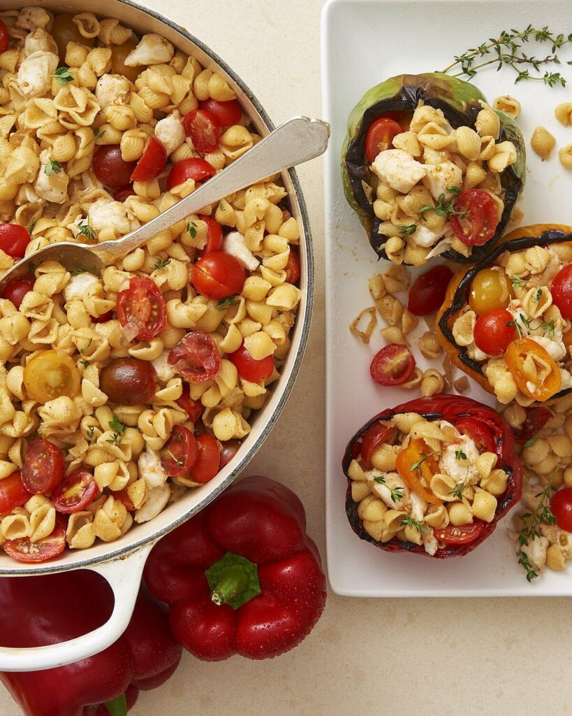 A white pot of Crab Pasta Salad with chunked tomatoes sits beside a white plate of grilled red pepper halves filled with the pasta salad.