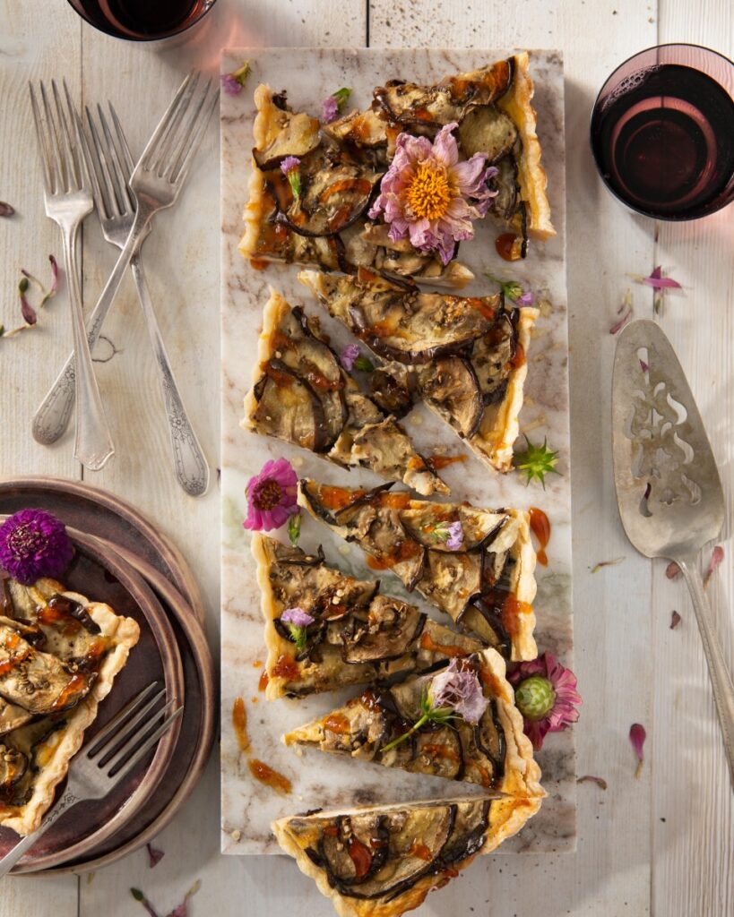 An eggplant and smoked mozzarella tart cut into pieces on a white table beside silverware and plates.