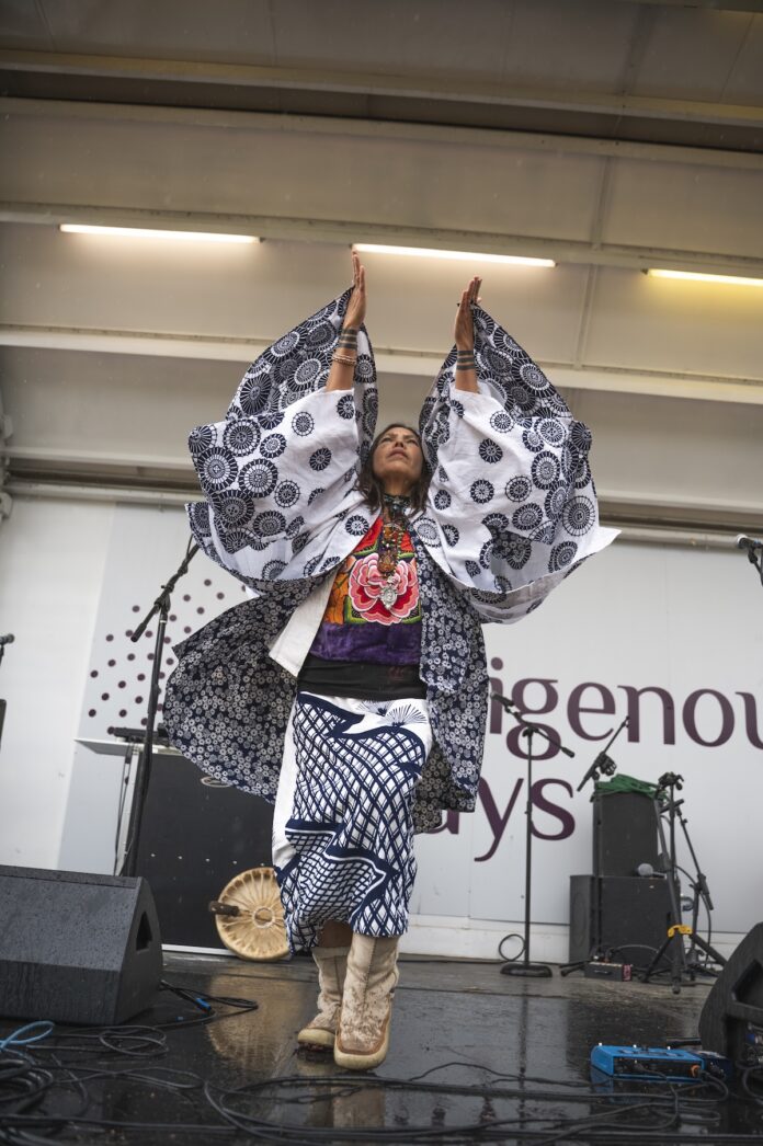 How IndigenousWays Is Preserving Native American Culture and Community in New Mexico -performer Madi Sato - pix by Mariano Frisoli - TABLE Magazine A performer on a stage with her arms raised above her head, and dressed in indigenous Native American attire at a festival.