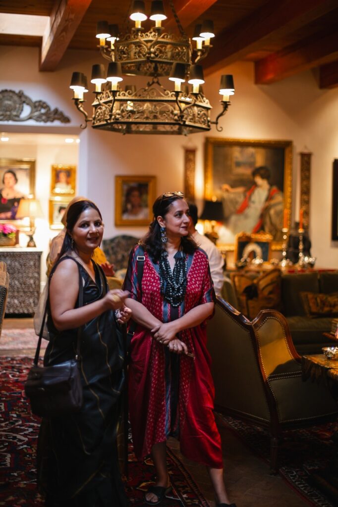 Two women in long dresses stand under a chandelier.