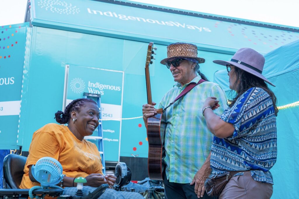 A man in a hat with a guitar talks to a woman sitting with a yellow shirt and another woman with a hat.