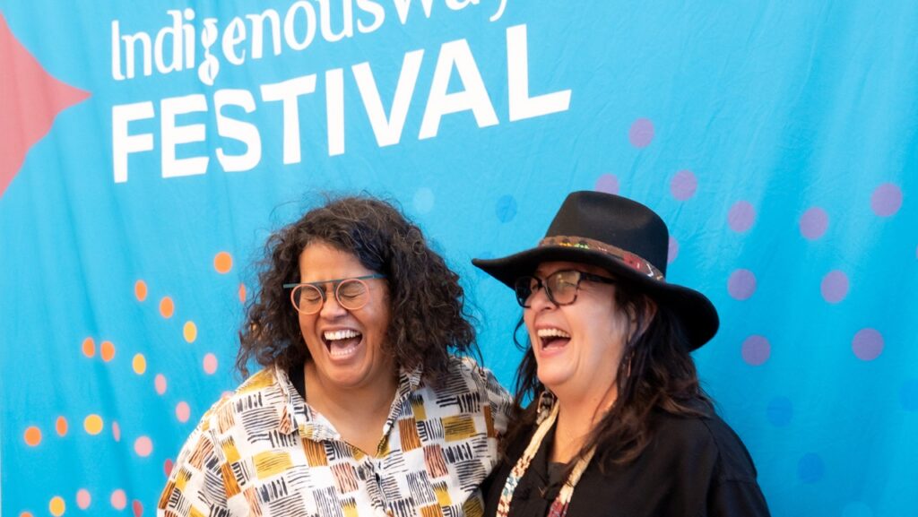 Two women laugh in front of the IndigenousWays Festival sign in blue.