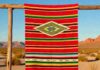 Shop New Mexico Artisan Fashion, Jewelry, and Home in Vibrant Orange A red striped New Mexico textile cloth hangs over a wood frame in the desert.