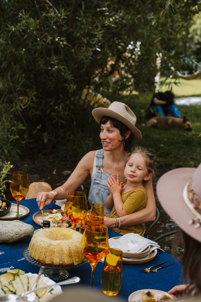A woman in a hat eats at the table with a child in her lap.