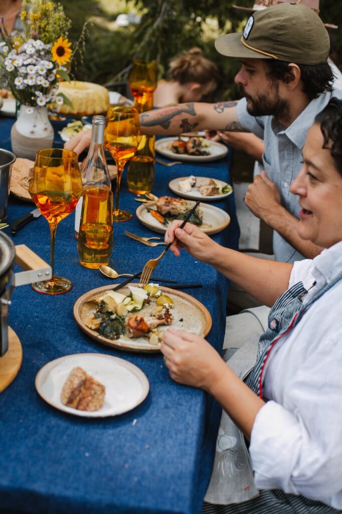A woman and a man eat from plates on a blue table with orange wine in front of them.