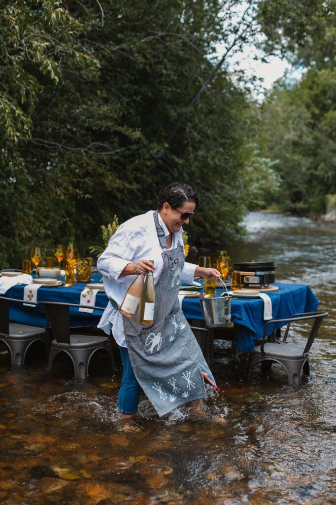 A woman walks in front of a table in a river carrying wine bottles.