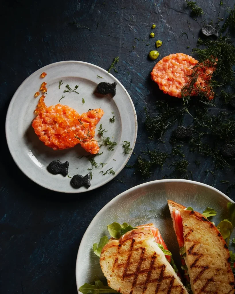 On a white plate to the left sits Salmon Tartare in the shape of a goldfish. To the right sits Salmon Grilled Cheese.