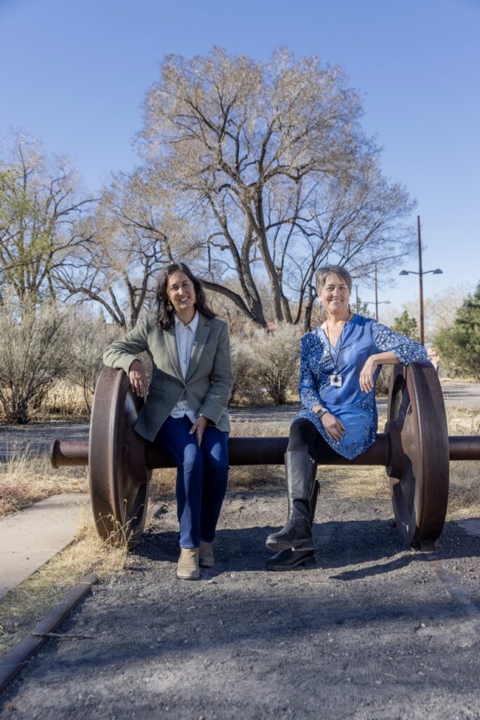 Two women sit on an old folk art bench outside.