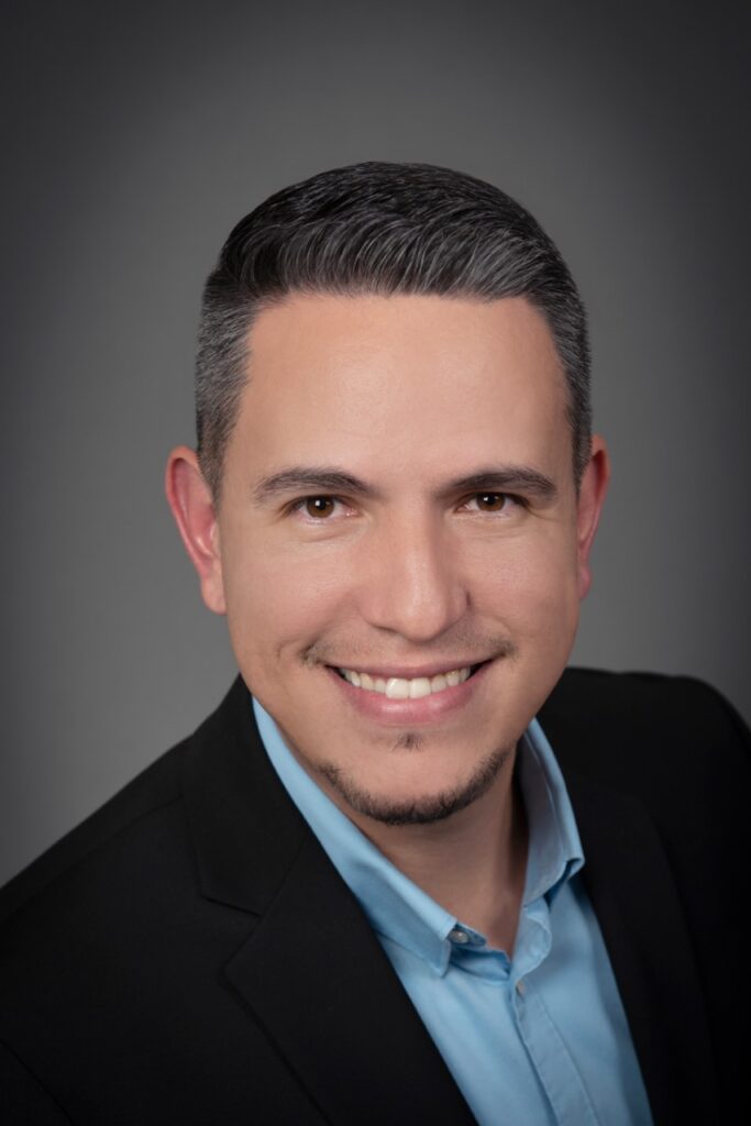 A portrait headshot of Marcos Zubia in a blue button up and suit jacket on a grey background.