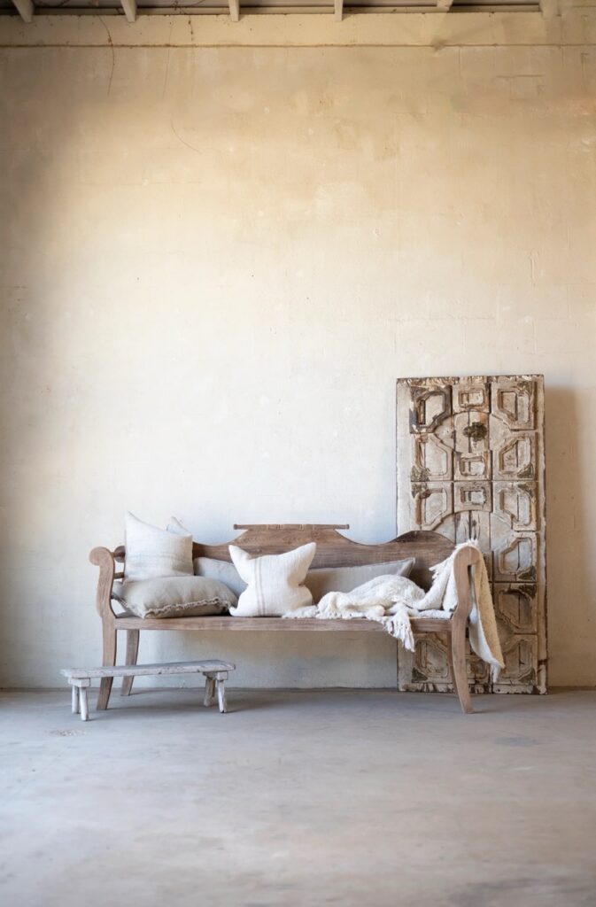 A wood bench sits against a beige wall with pillows on it and a cabinet to the right. 