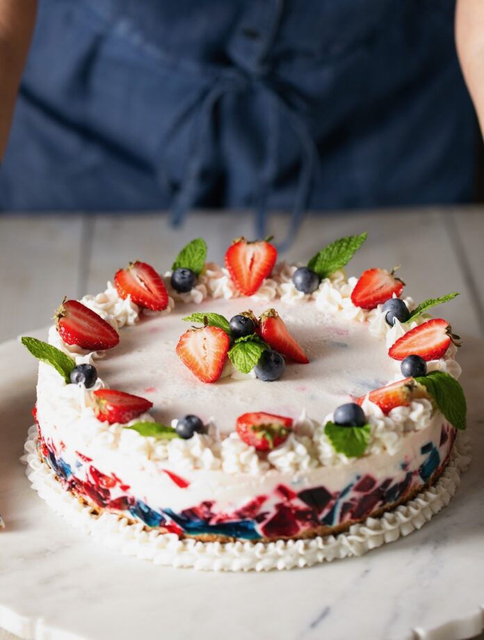 Red White and Blue Stained Glass Gelatin Cake A full gelatin cake in red, white, and blue colors decorated in strawberries, blueberries, and mint sits on a picnic like table.