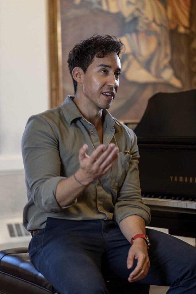 A man sits next to a piano using his hand as he talks.