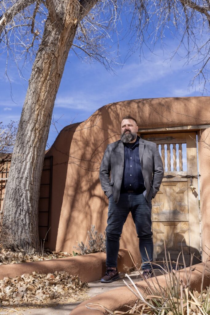 A man in a jacket and jeans stands outside a historical New Mexico site outdoors.
