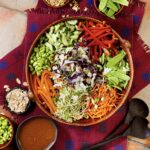 A bowl full of a salad with colorful crunchy vegetables in an Asian salad beside small bowls of veggies and a bowl of dressing.