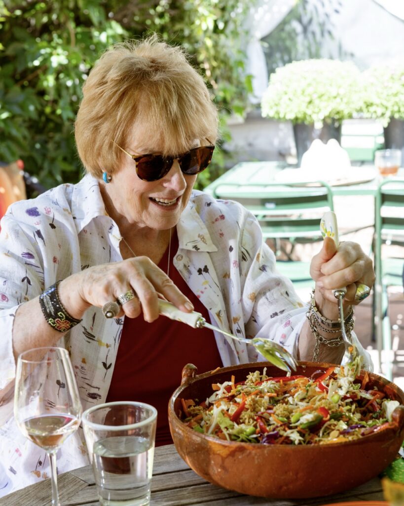 A woman in a white jacket and sunglasses tosses a salad at a table.