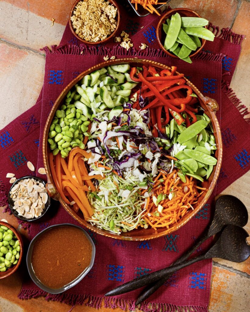 A bowl full of a salad with colorful crunchy vegetables in an Asian salad beside small bowls of veggies and a bowl of dressing.