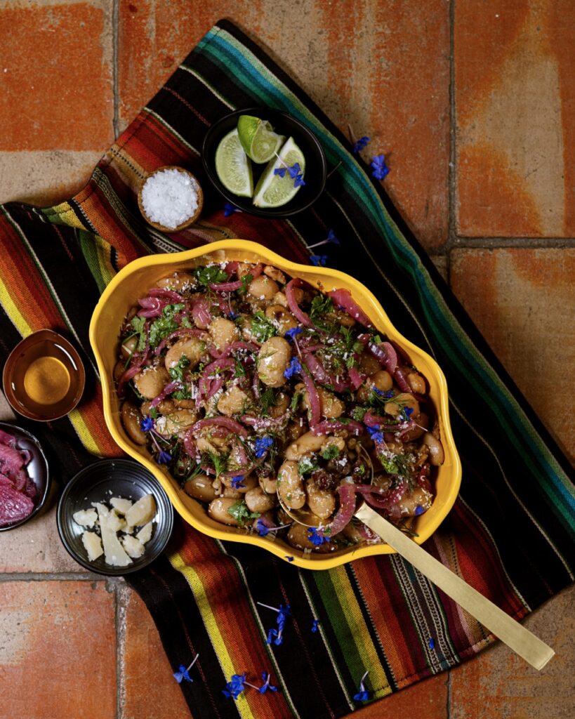 A yellow swiggly bowl with a Bean Salad inside beside little dishes of spices and limes.