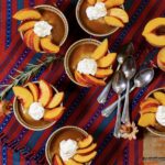 Small tins of Butterscotch Budino with whipped cream and slices of peaches on top, all on a red and blue cloth.