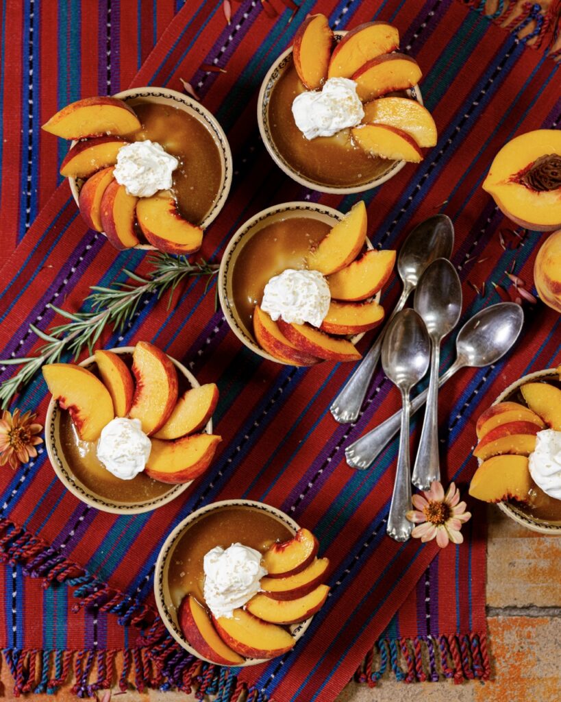 Small tins of Butterscotch Budino with whipped cream and slices of peaches on top, all on a red and blue cloth.