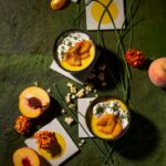 Two bowls of corn custard topped with pickled peaches and whipped goat cheese on a green textured background with fresh peaches and corn kernels.